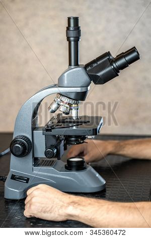 Male Laboratory Assistant Examining Biomaterial Samples In A Microscope. Cllose Up Hands In Blue Rub