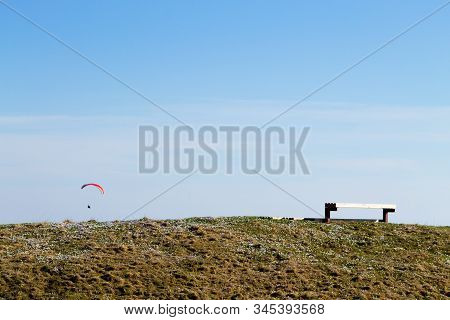 Paraglider On Blue Sky With Empty Bench On Foreground. Borso Del Grappa, Famous Paragliding Site, It