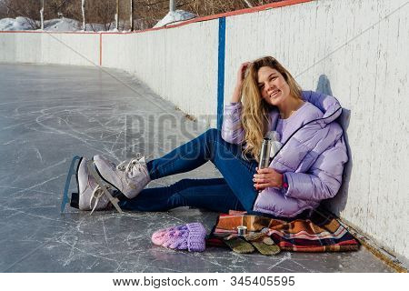 Lovely Young Woman Relaxing After Riding Ice Skates And Drinking Hot Drink From Termo Pot On The Ice