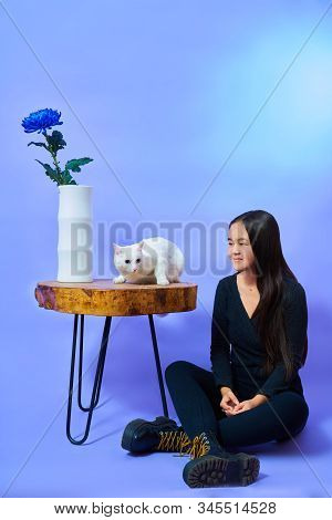 A Girl Of Asian Appearance Sits On The Floor Next To A Wooden Table - On The Table Sits A White Cat 
