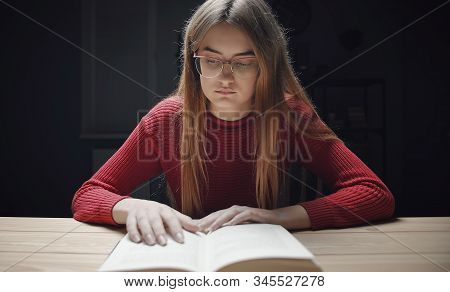 Focused Girl In Glasses Reading Book Following Text By Hand Against Dark Background