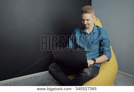 Smiling Guy Working On Laptop Holding It On His Lap While Sitting On Yellow Bean Bag Chair