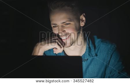 Young Bearded Man Smiling Whilst Using His Laptop At Night, Concept Of Modern Technology Use
