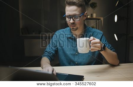 Serious Young Man Holding Cup Having Coffee Sitting At Table Looking At Pc In Dark Late Night Indoor