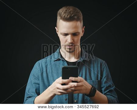 Front View Of Serious Young Man In Jean Shirt Typing Message On Phone, Isolated Black Background