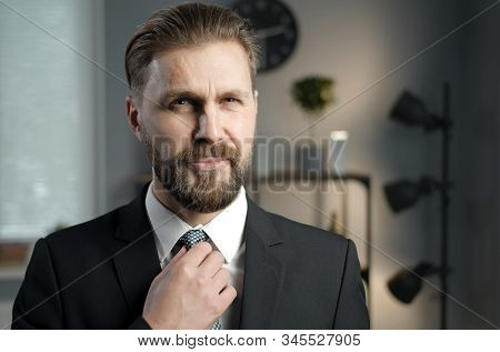 Bearded Mature Businessman In Black Formal Suit Fixing His Check Necktie Looking At Camera