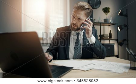Bearded Mature Businessman In Classic Wear Talking On Smartphone While Looking Laptop Screen In Offi