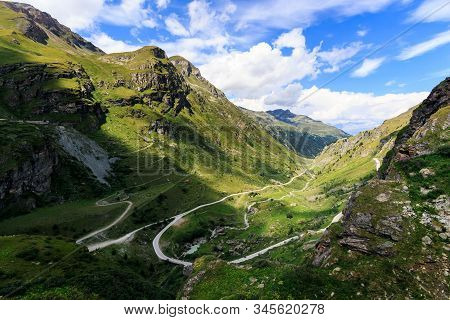 Panoramic View Of A Valley In The Pennine Alps With A Winding Road, Green Alpine Pastures Under A Bl