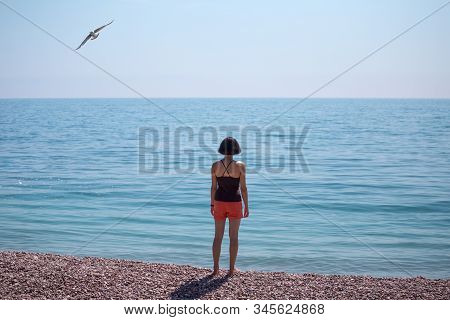 A Woman Stands On The Seashore And Looks Into The Distance. Slender Girl Looks At The Sea. A Brunett