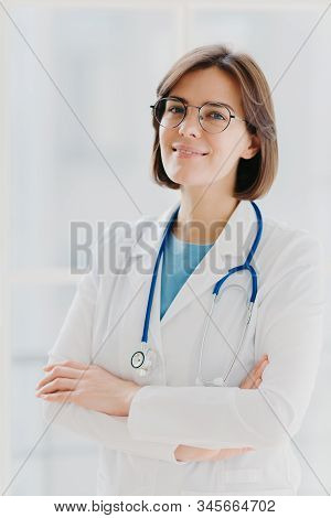 Shot Of Confident Brunette Female Doctor Or Surgeon Stands With Hands Crossed, Wears White Uniform, 