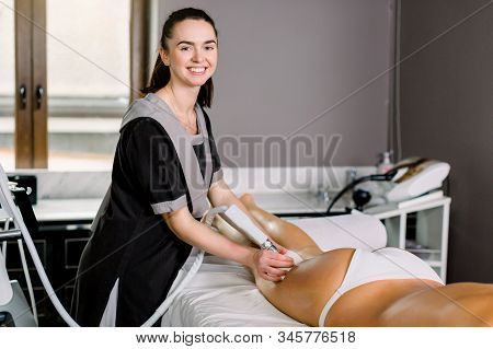 Young Smiling Woman Cosmetologist With Roller Doing Buttocks Massage For Her Female Client In Modern