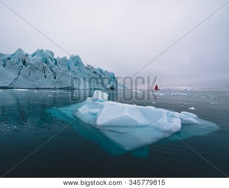 Beautiful Red Sailboat In The Arctic Next To A Massive Iceberg Showing The Scale. Cruising Among Flo