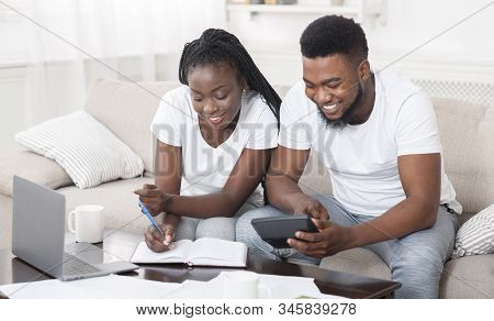 Young Happy African American Couple Planning Family Budget Together, Sitting On Sofa At Home, Using 
