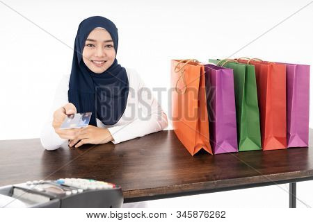 Confident happy smiling female muslim woman pay for her shopping with credit card with EDC machine using as consumerism and shopaholic concept. Studio shot on white background.