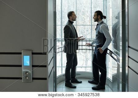 Two elegant businessmen discussing working points or terms of agreement inside elevator of large business center