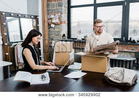 Content young clothes production employees packing sewed clothes in boxes while sorting it for sale in studio