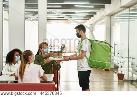 Male Employee Taking Pizza And Coffee From Courrier During Lunch With Female Colleagues At Work. Peo