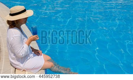 Luxury Senior Woman Dangling Her Legs In An Outdoor Swimming Pool Holding A Blue Refreshing Cocktail