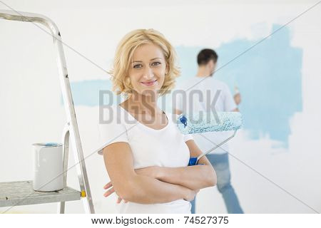 Portrait of woman holding paint roller with man painting wall in background