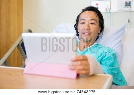 Mature male patient using digital tablet while reclining on bed in hospital