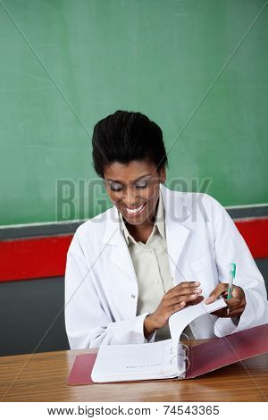 Happy young female teacher reading paper in binder at desk in classroom