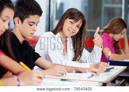Portrait of confident teenage girl gesturing thumbs up while friends studying in classroom