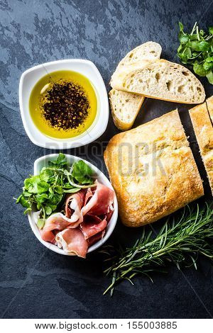 Bread ciabatta, jamon ham serrano paleta iberica, arugula, olive pepper oil, rosemary and glass of red wine on stone slate black background. Top view copy space