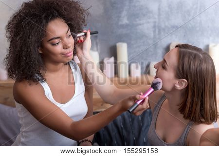 May I help you. Positive smiling involved girls putting the makeup on each other and sitting on the bed while getting ready for a party