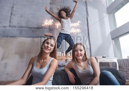 Wonderful home party. Amused smiling young girl jumping on the bed in the bedroom while raising her hands and expressing joy with friends
