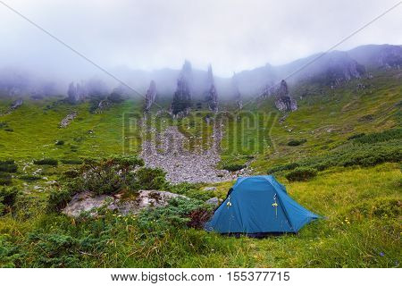 A solitary tourist tent stands on a meadow and around headlong rocky mountains and dense fog on a summer morning.