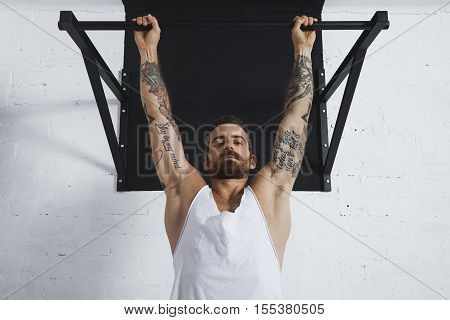 Strong tattooed in white unlabeled tank t-shirt male athlete shows calisthenic moves close up of classic pullup Hanging on pull bar