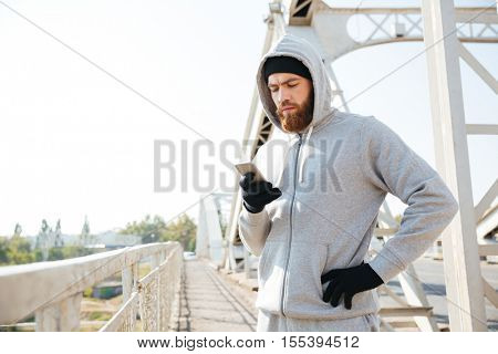 Concentated bearded sports man typing on mobile phone while standing outdoors at the bridge