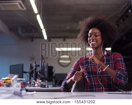 black woman in modern office at her workplace speeking on phone over earphones and working on laptop computer