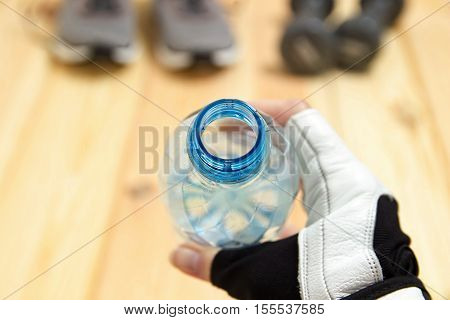 Bottle of water in hands close-up, cropped view, foreground, Sneakers and dumbbells on a wooden background, sport and Fitness, drinking from the bottle
