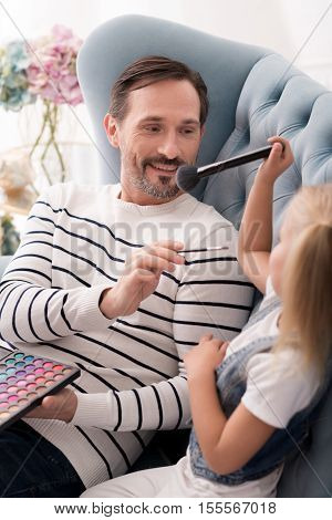 Playing with an eye shadow palette. Nice cheerful optimistic man holding a brush and smiling while putting make up on his daughter