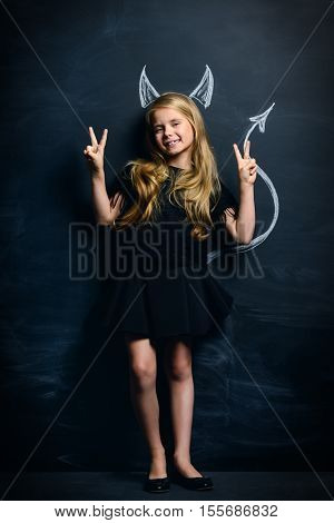 Funny little imp girl. Cute child girl posing with imp horns and tail drawn on a blackboard.