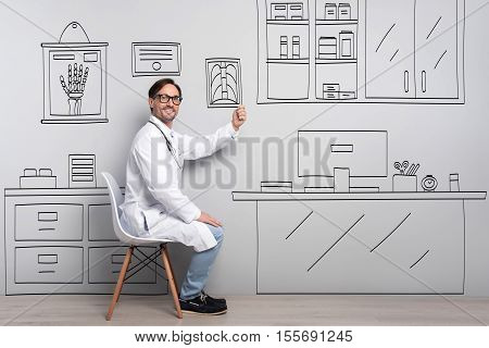 Job like a hobby. Glad handsome young man smiling and holding roentgenogram while sitting by the table.