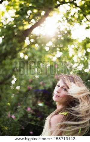 Beautiful young woman with long blowing blonde hair against green tree. Sunny summer day.