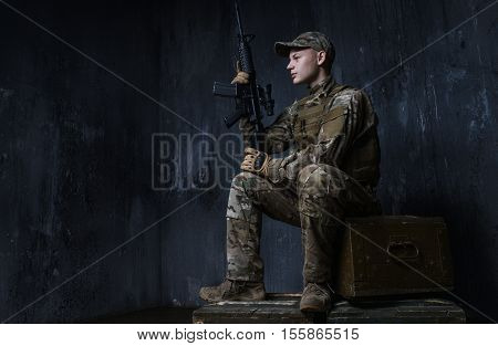 Young soldier sitting on crates of rifle and confidently looks ahead. Around the world those guys keep us safe