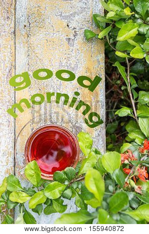 Glass cup of tea on a wooden surface, green grass around. Red orange flowers. Text Good morning on the background.