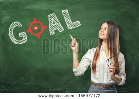 A young woman holding her glasses and pointing the finger at the word goal written on the green chalkboard wall. Set a target and and keep trying until you reach it. Dedication in discharging important tasks. To face the future.