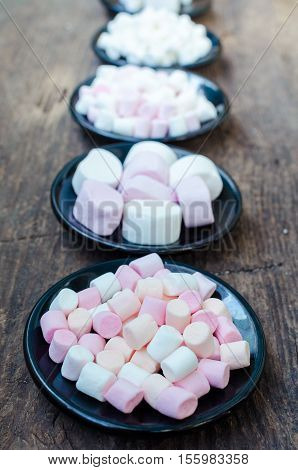 Colorful mini marshmallows in a black plates on a wooden background. Different mini white pink and orange puffy marshmallows. Marshmallow concept.