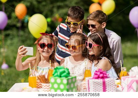 holidays, childhood and technology concept - happy kids in sunglasses taking selfie by smartphone on birthday party at summer garden