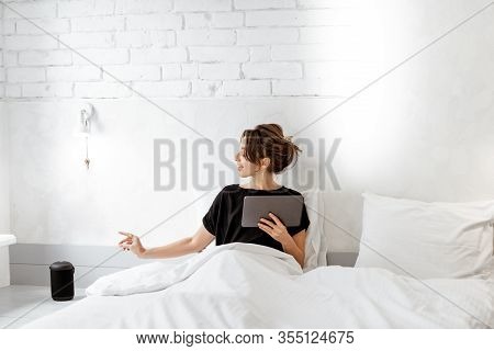 Young Woman Lying In Bed With A Digital Tablet And Smart Speaker, Controlling Home Devices With A Vo