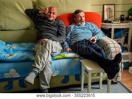 Two bored men are sitting on the couch in front of the TV at home, real people every day life