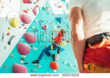 Father And Teenager Son At Indoor Climbing Wall Hall. Boy Is Hanging On The Rope Using A Climbing Ha