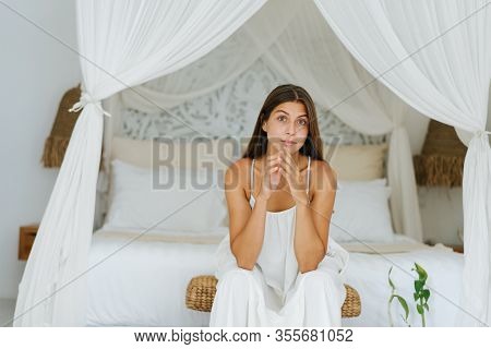 Woman With Stepeled Hands In White Dress Is Sitting Next To A Luxury Double Bed