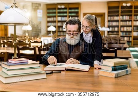 Elderly Man Grandfather And His Granddaughter Reading Exciting Book Together In Vintage Old Library,