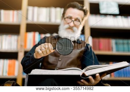 Handsome Smiling Senior Bearded Man, Librarian Or Professor, In The Library, Sitting On The Backgrou