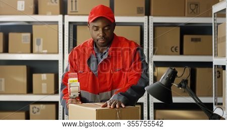 Young African American Postal Male Worker In Uniform Packing Carton Box At Delievery Department In P